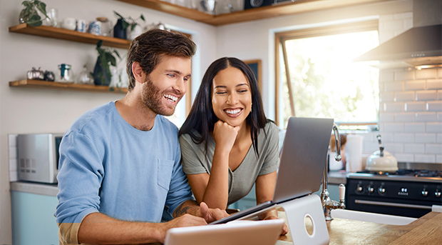 A happy couple sits in their kitchen on their laptop looking at their savings.